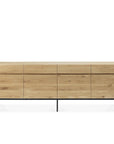 The Ethnicraft Ligna Sideboard features four drawers and four doors in European oak with a light natural finish, set on slim black metal legs, shown against a white background.