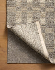 A beige and gray checkered rug is partially folded to show a Loloi Cushion Grip Rug Pad - 3/8, made from recycled synthetic fibers, on a light wood floor—perfect for use on both hard surfaces and carpet.