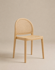 The Luk Dining Chair by Wooden Stories features a light wooden arch silhouette, rounded backrest, and woven cane seat. Set against a beige background, its minimalist design and rattan details add modern, contract-grade style.