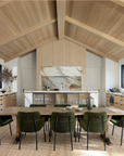 A modern kitchen and dining area features a vaulted wood ceiling, light wood cabinets, marble backsplash, and the SOBU Mars Live Edge Dining Table in white ash with brass T legs, surrounded by eight green velvet chairs and large windows.
