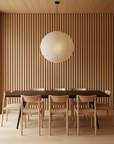 A minimalist dining room features a black rectangular table, six Wooden Stories Noki Dining Chairs in Light Oak, a slatted wood wall, light wood floors and ceiling, and a large round paper lantern pendant above the table.