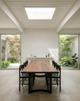 A minimalist dining room features SOBU's Mars Live Edge Dining Table in walnut with curved legs, paired with black chairs. Skylight and sliding glass doors reveal garden views, while neutral tones and natural light create an airy, modern vibe.