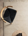 Overhead view of the Armadillo&Co Malawi Rug - Chino, a hand-knotted beige rug with fringed edges, beside a rounded wooden chair with a black woven seat and a light brown circular table for an artisanal touch.