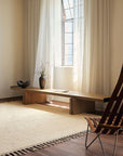 A minimalist room features Armadillo&Cos Malawi Rug in Chino with fringe edges, a wooden bench below a tall window with sheer curtains, a wooden chair, and a dark vase with dried grass. Sunlight streams in, creating an artisanal feel.