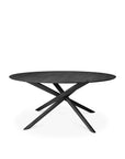 The Ethnicraft Mikado Dining Table Round in black oak features a smooth top and four diagonally crossing legs forming a sculptural base. Crafted from European oak with a black varnished finish, it is shown on a plain white background.
