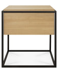 The Ethnicraft Monolit Nightstand - Oak features a light wood drawer and top crafted from European oak, supported by a black metal open frame base. This modern piece embodies classic European refinement and is shown against a white background.