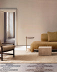 A modern living room with a tan sofa, armchair, and neutral rug features the Wooden Stories Troubadour Lounge Chair in fabric and a cube coffee table. Sunlight pours through a large window, with an open door revealing another bright room beyond.
