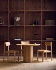 A round wooden table with two Wooden Stories Noki Dining Chairs in Light Oak sits before a floor-to-ceiling wooden shelving unit displaying books, a skull model, and a camera.