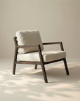The Nysse Lounge Chair | Fabric by Wooden Stories features light beige cushions and a dark wooden frame, set on a pale floor with soft natural light gently casting shadows nearby.