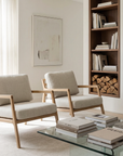 A minimalist living room features two Wooden Stories Nysse Lounge Chairs in leather, a glass coffee table with art books, built-in shelving for books and firewood, and a large abstract artwork. Natural light emphasizes the Scandinavian style.