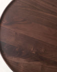 A close-up view of the Bystander Side Table by Wooden Stories, featuring a round, dark wood tabletop with a smooth finish and visible natural grain patterns.