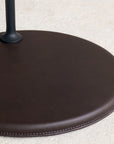 A close-up of the Wooden Stories Bystander Side Table shows its round, dark brown leather base with visible stitching on a light-textured surface and a black metal pole attached at the top left.