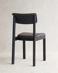The Noki Dining Chair - Black Oak - Leather by Wooden Stories, featuring a minimalist Japanese-inspired design with a curved back and padded seat, is set on a light textured floor against a neutral wall, facing away from the camera.
