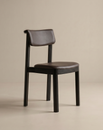 The Noki Dining Chair | Leather by Wooden Stories is a modern solid wood chair featuring a dark frame, brown leather cushioned seat, and curved padded backrest, shown against a plain beige background. Contract grade.