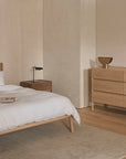 Minimalist bedroom featuring a wooden bed frame, crisp white bedding, a brown accent pillow, wooden nightstand, black lamp, and the Nell Tall Dresser by Wooden Stories with premium metal runners for smooth bedroom storage solutions. Decor accents and a beige rug on hardwood floors complement the neutral tones and simple, modern design inspired by the Danish Collection.
