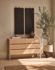 The Nell Tall Dresser by Wooden Stories, crafted in light wood with premium metal runners and accents, pairs beautifully with two dark wall panels, a potted tree, and sunlight streaming in to create a serene minimalist interior of neutral tones—an elegant expression of the Danish Collection approach to bedroom storage.