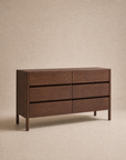 The Nell Wide Dresser by Wooden Stories is a modern dark wood piece from the Danish Collection, featuring six drawers and set against a plain beige background. Its simple rectangular silhouette showcases clean lines with no visible handles and premium metal runners for seamless drawer operation—making it an ideal choice for stylish bedroom storage.