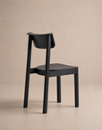 The Noki Dining Chair | Wood by Wooden Stories features a modern, minimalist solid wood design with a curved backrest, shown from behind at an angle against a plain beige background.