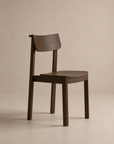 The Wooden Stories Noki Dining Chair | Wood is a minimalist solid wood chair with a curved backrest and seat, dark brown finish, and clean lines. Contract grade quality ensures lasting durability. Shown against a plain beige background.