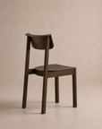 The Noki Dining Chair | Wood by Wooden Stories is a contract-grade solid wood chair with a flat seat and curved backrest, displayed from behind at an angle against a neutral beige background.