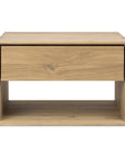 The Ethnicraft Nordic II Bedside Table is crafted from solid European oak with a natural finish, featuring a central drawer and open shelf, shown against a white background.