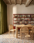 A sunlit room with a terrazzo floor features the Oli Dining Table by Wooden Stories and cane chairs. A red metal bookshelf against the wall displays books and decor, while green curtains frame the window and wooden beams cross the ceiling.