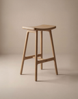 The Odd Counter Stool | Wood by Wooden Stories is a minimalist solid oak stool with a curved seat, four legs, and visible natural wood grain, shown against a neutral beige background. Its simple, modern design makes it perfect for any space.