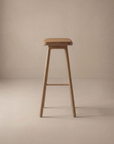 The Odd Counter Stool | Leather by Wooden Stories features a minimalist solid oak frame with a brown leather cushioned seat, shown on a light beige background—ideal for stylish and sturdy kitchen island seating.