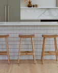 Modern kitchen with a white marble island and three Wooden Stories Odd Counter Stools in light oak with leather seats, plus gold faucet and minimalist Danish decor featuring wooden bowls and neutral vases in natural materials.