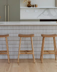 A modern kitchen with a white island featuring vertical tile lines, minimalist cabinetry, a gold faucet, and three Wooden Stories Odd Counter Stools | Leather. Light wood flooring and decorative accents complete the look.