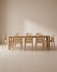 A minimalist dining room showcases the Wooden Stories Oli Dining Table in solid American oak with rounded edges, paired with eight matching wooden chairs featuring woven cane backs, set against a neutral beige background.
