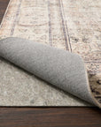 A corner of a patterned area rug is lifted to show the Loloi Cushion Grip Rug Pad - 1/8, made from recycled synthetic fibers, on a dark wooden floor.