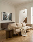 A neutral living room featuring the Wooden Stories Pensive Sofa | Fabric, chunky knit blanket, wooden side table with mug, framed artwork, wooden console, large window, and arched doorway to a staircase.