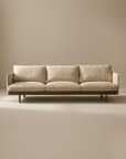 A minimalist beige three-seater Pensive Sofa | Fabric by Wooden Stories, with plush cushions and a wooden base inspired by mid-century design, is set against a plain, light-toned background.
