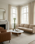 A bright, elegant living room with tall windows, a light beige sofa, brown leather chairs, and the Wooden Stories Eden Coffee Table in wood and stone. Beige curtains and minimalist decor evoke Scandinavian simplicity.