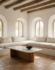 Minimalist living room with white built-in sofas, arched windows, wooden beams, solid oak accents, terracotta tile floor, and a Wooden Stories Pivot Coffee Table (Rectangular - Stone & Wood) topped with a black bowl. Sunlight softly fills the space.