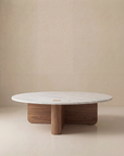 The Wooden Stories Pivot Coffee Table | Round—Wood & Stone features a white marble top and solid oak intersecting base, displayed against a plain beige background.