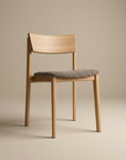 The Poise Dining Chair | Light Oak with Fabric by Wooden Stories features a light brown oak frame, curved backrest, and gray cushioned seat—perfect for contract grade spaces, shown against a plain beige background.