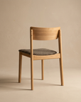 The Poise Dining Chair by Wooden Stories, in light oak with a cushioned gray fabric seat and curved backrest, is shown from behind and at an angle on a beige surface with a matching background.