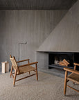 A minimalist living room with a large concrete fireplace, two woven wood armchairs on the Pebble Rug - Whippet by Armadillo&Co, a small round side table, and a slim black floor lamp against textured gray walls and ceiling.