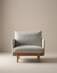The Pensive Lounge Chair | Fabric by Wooden Stories, with light gray upholstery and a wooden frame inspired by mid-century Danish design, stands out against a plain beige background.