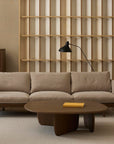 A minimalist living room with a beige Pensive Sofa by Wooden Stories, round wooden coffee table with a yellow book, side table, dark wood cabinet, modern black lamp, and light wood slatted wall panel.