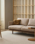 A minimalist living room featuring the Pensive Sofa by Wooden Stories, a wooden armchair, small round side table, light rug, and empty wooden shelves against the wall, softly lit by natural light from tall windows.