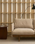 A modern living room featuring the customizable Pensive Sofa | Fabric by Wooden Stories in beige, a dark wooden side table with a book, a black adjustable wall lamp, and a wooden slatted accent wall in the background.
