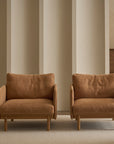 Two tan Pensive Lounge Chairs by Wooden Stories sit side by side on a light rug next to a small dark wood side table, with two tall wooden cabinets against a beige vertically paneled wall in the background.