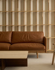 Minimalist living room featuring the Wooden Stories Pensive Sofa in brown leather, a light wood coffee table, beige armchair, and a wooden slat wall. Neutral colors and clean lines create an inviting atmosphere.