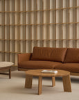 A minimalist living room with the Wooden Stories Pensive Sofa in brown leather, a light wood round coffee table with books, a beige cushioned bench, and a decorative wooden grid wall in the background.
