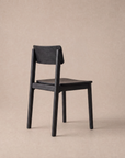 The Poise Dining Chair | Wood by Wooden Stories, featuring a simple modern design in dark solid oak from the Danish Collection, is positioned on a light beige background, angled slightly away to display its backrest and seat.