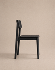 The Poise Dining Chair | Wood by Wooden Stories is shown in profile against a plain beige background. This minimalist black solid oak chair features a simple, modern design with straight legs and an angled backrest, making it contract grade and suitable for both residential and commercial spaces.