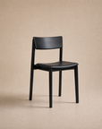The Poise Dining Chair | Leather by Wooden Stories, featuring a simple black solid oak frame with a curved seat and backrest, stands on a neutral beige background, exemplifying the refined craftsmanship of the Wooden Stories collection.